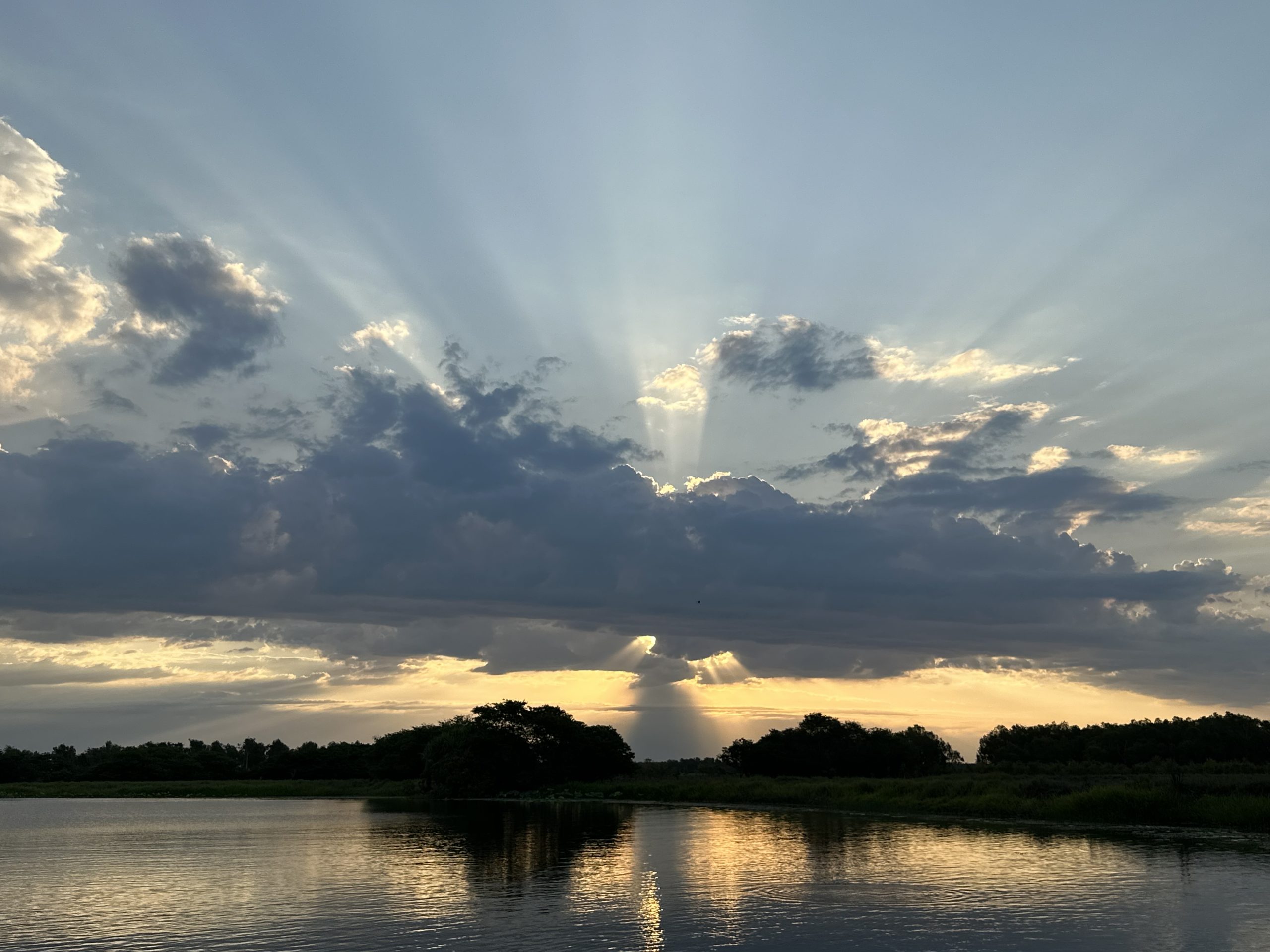 Sun breaking through clouds over a tranquil lake at dusk